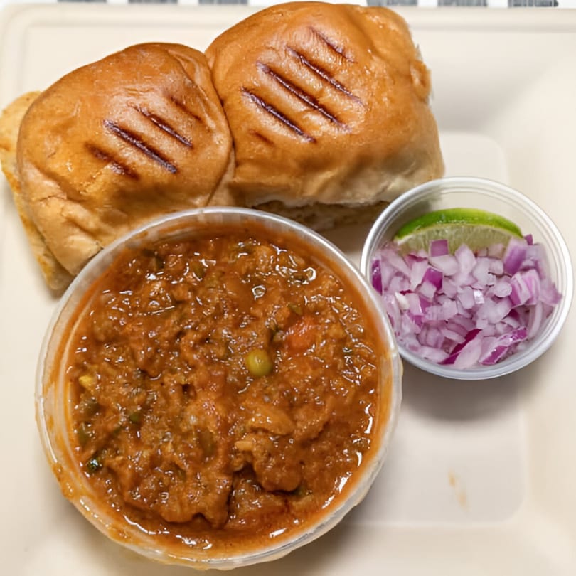 Desi Chowrastha's Pav Bhaji with grilled buns, a bowl of vegetable curry, and a side of chopped red onions and lime.