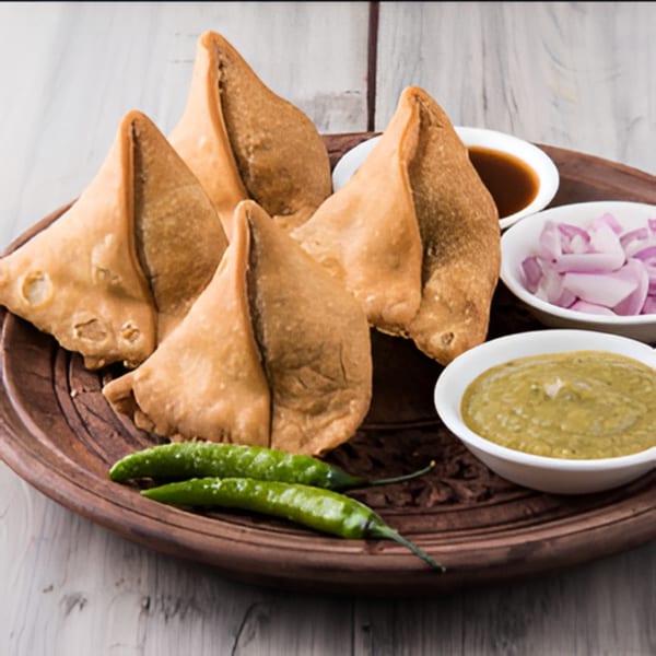 Four golden-brown samosas on a wooden plate with green chilies, red onions, and two dipping sauces.