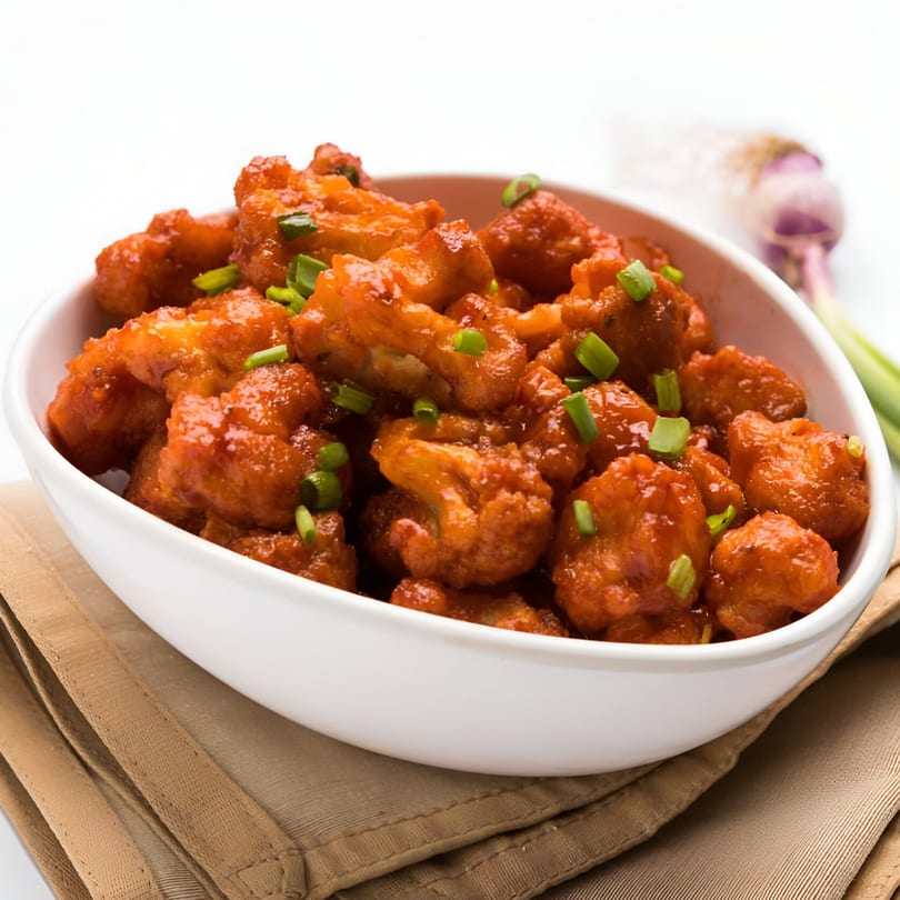 Desi Chowrastha's Gobi Manchurian, a bowl of fried cauliflower florets in a spicy red sauce, garnished with green onions.