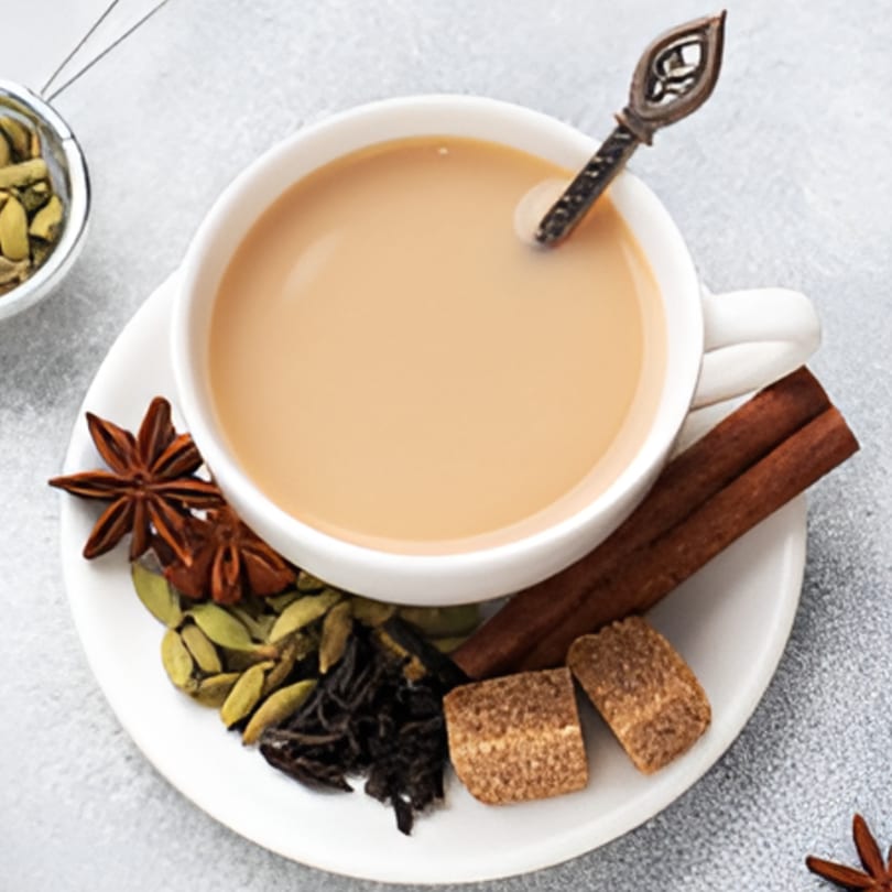 A cup of Indian chai with a decorative spoon, surrounded by star anise, cardamom, cinnamon, and sugar cubes.